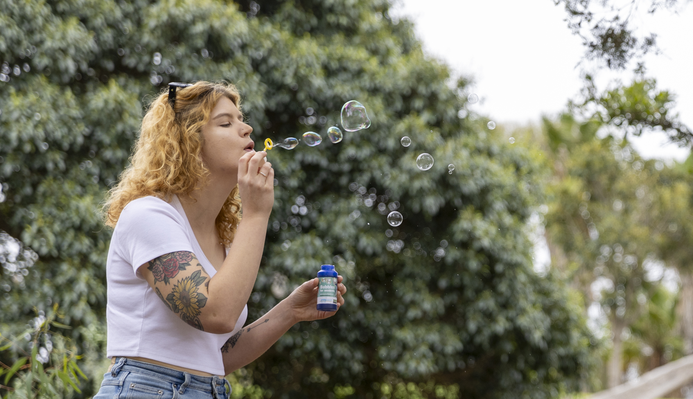 A woman blows bubbles with a bubble wand, there are green trees in the background