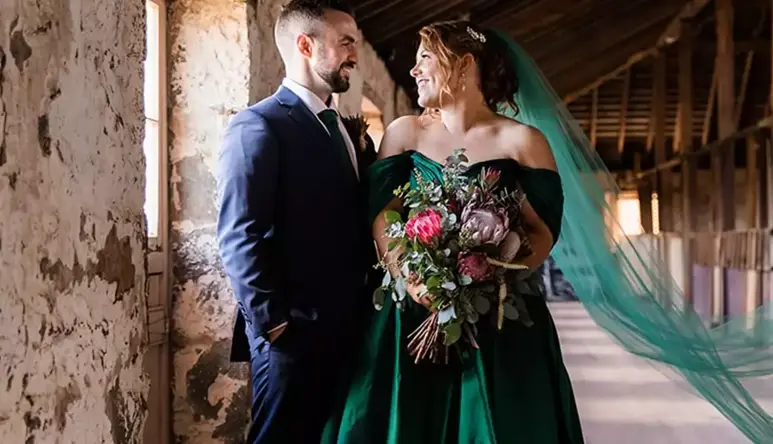 A man and woman dressed in formal wedding wear are standing in a rustic room, looking at each other.