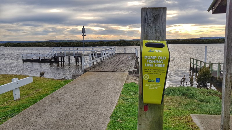A bin on a wooden pole in front of a jetty over an inland waterway; the bin has text on it that reads "Dump old fishing line here"