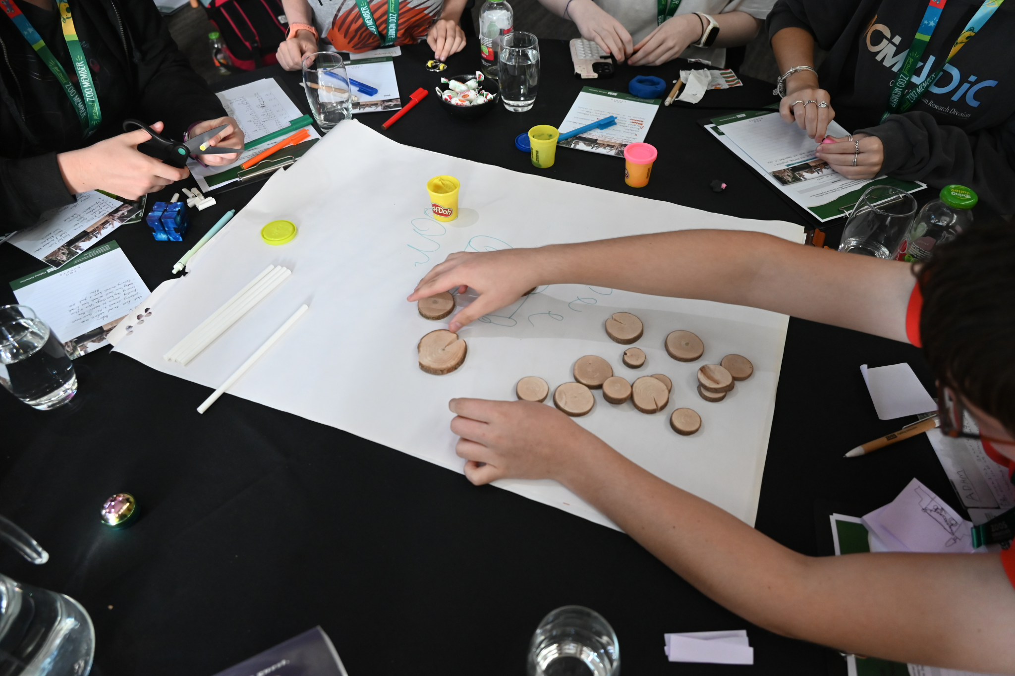 Five guests gathered around a black table, all engaged in a craft activity with paper, scissors, markers, three tubs of modelling clay and fifteen small wooden discs 