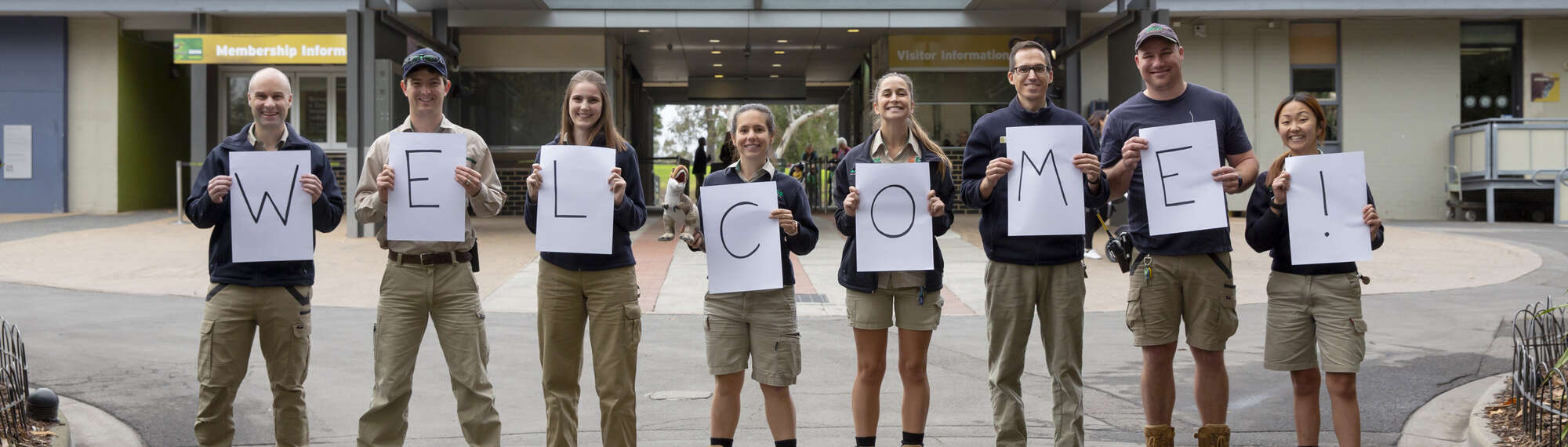 Eight Zoo staff each holding signs that spell out "W-E-L-C-O-M-E-(exclamation point!)."