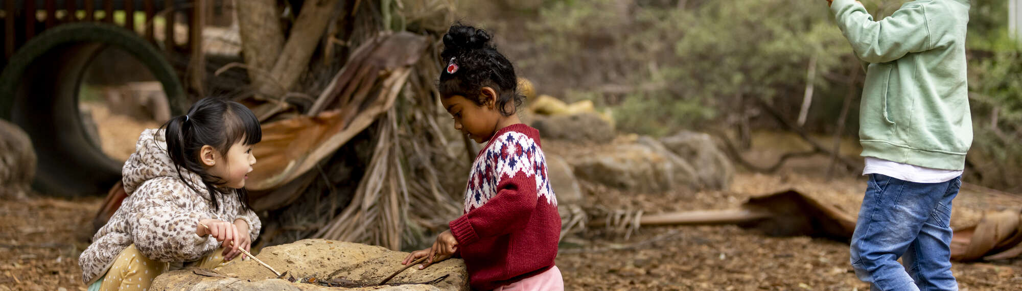 Three children playing with sticks and rocks in the outdoor playground.