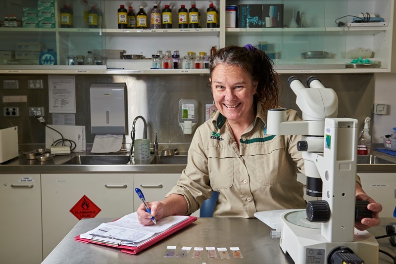 A smiling Vet, dressed in khaki, sits in front of a microscope with a clipboard and pen in her hand.