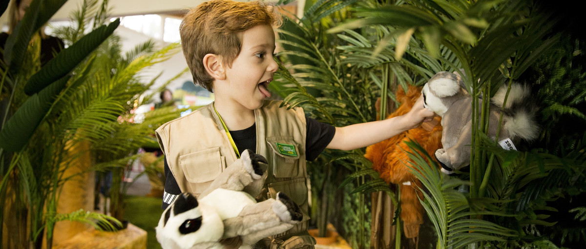 A young guest in a Keeper vest excitedly plays with two plush Lemurs, with an Orangutan and ferns behind him.