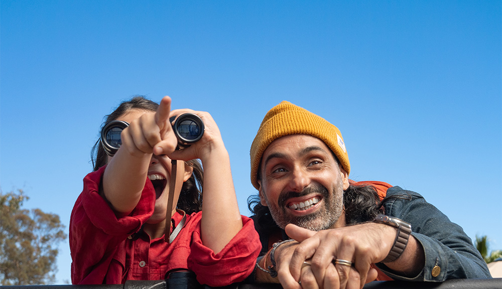 Two guests watching Elephants, the daughter excitedly pointing while looking though binoculars, with her dad looking on and smiling.