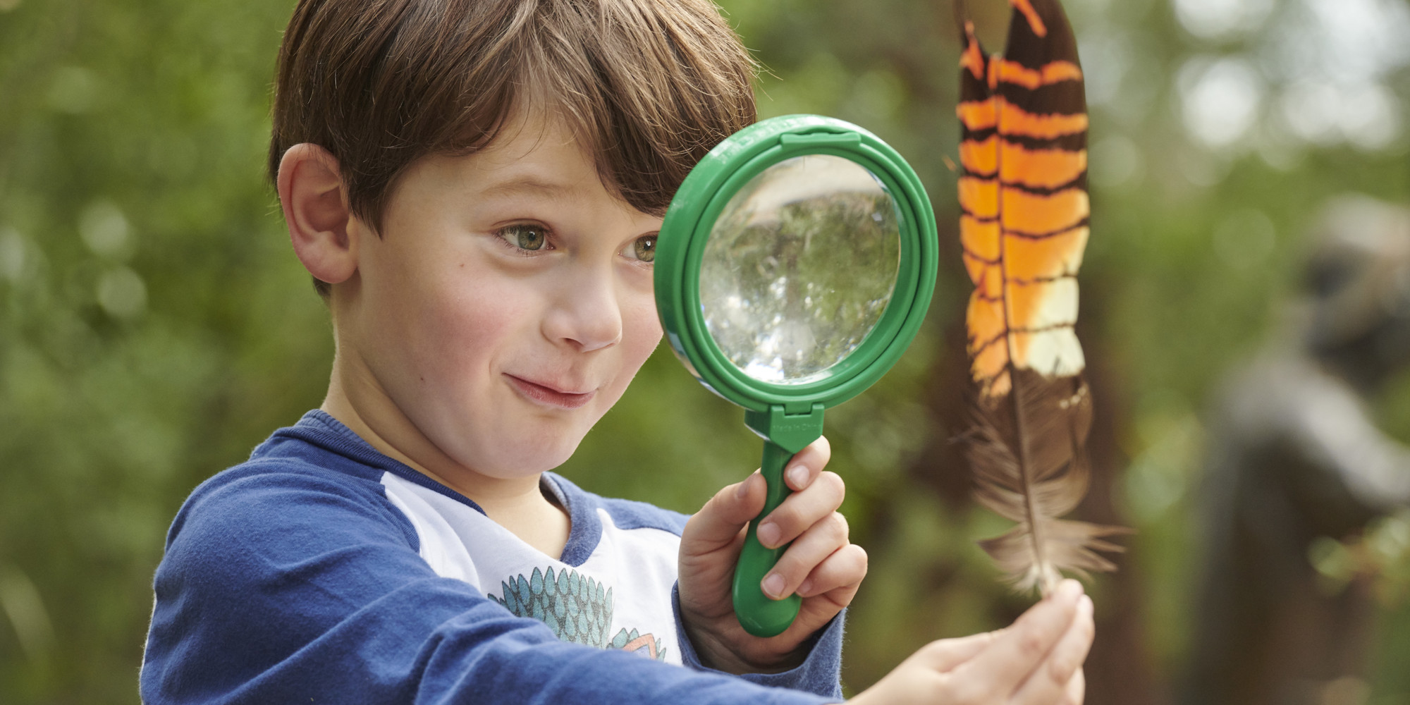 Kinder Keeper Kid examines a brown-and-orange feather with a green magnifying glass.