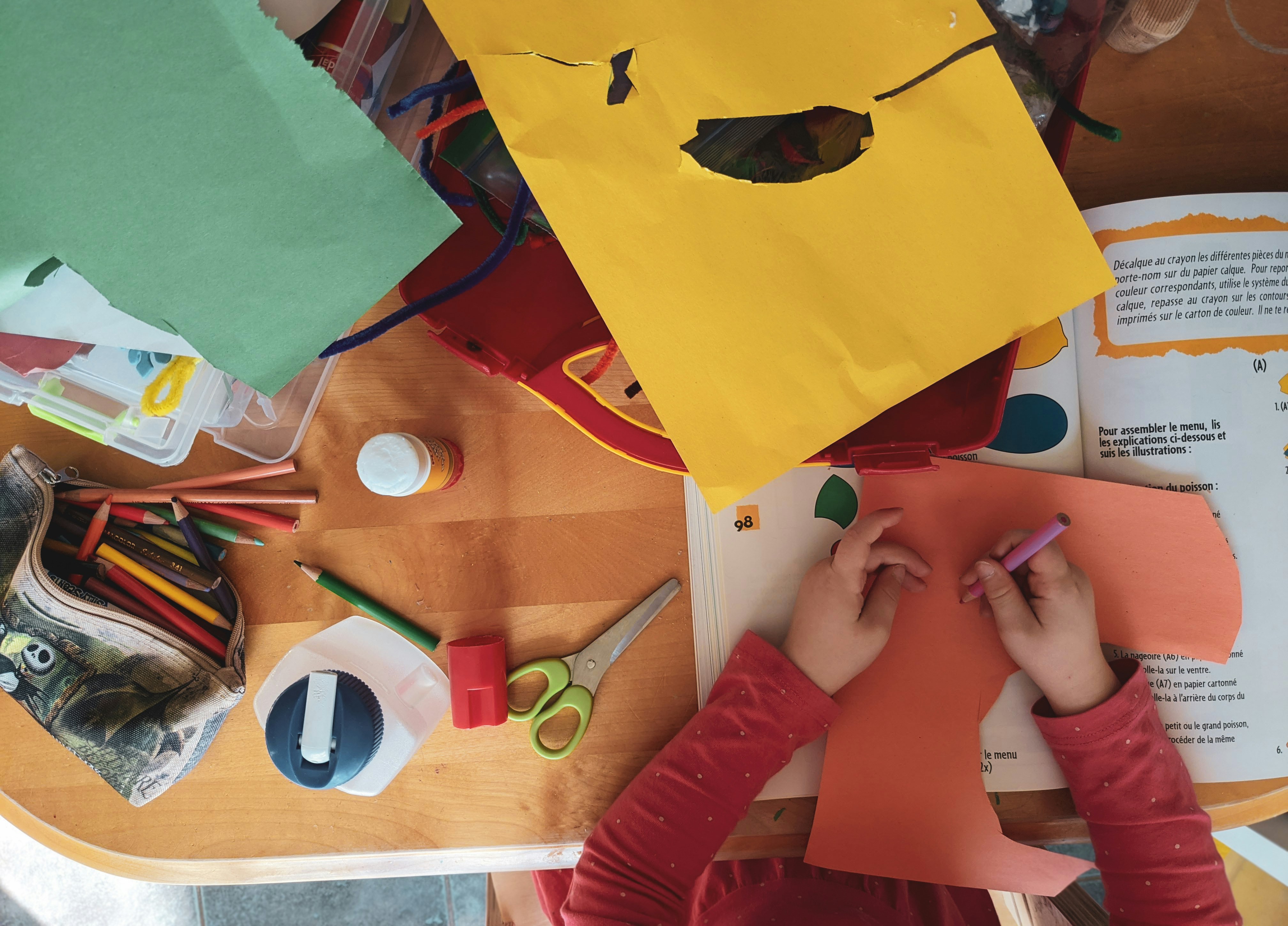 A top-down view of a child's desk featuring assorted coloured paper, pencils, glue, a bottle, scissors, felt wire, a sharpener and a (French) papercraft activity book.