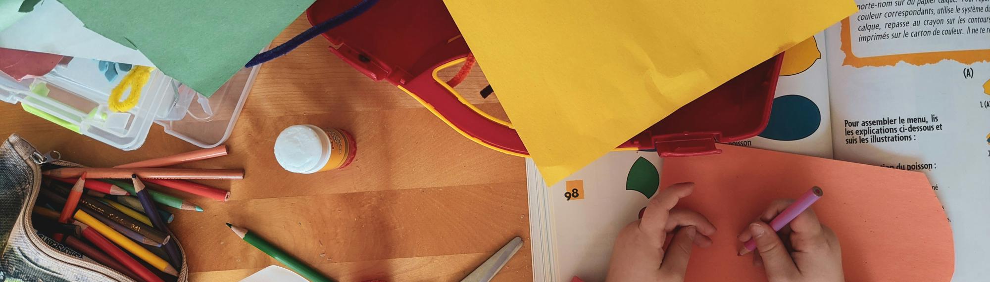 A top-down view of a child's desk featuring assorted coloured paper, pencils, glue, a bottle, scissors, felt wire, a sharpener and a (French) papercraft activity book.